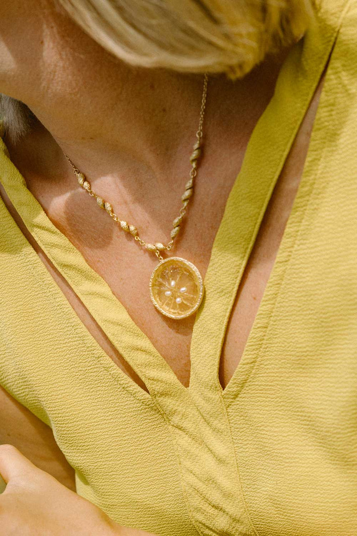 A close-up of a woman wearing a gold necklace with a lemon slice pendant, paired with a textured yellow top.