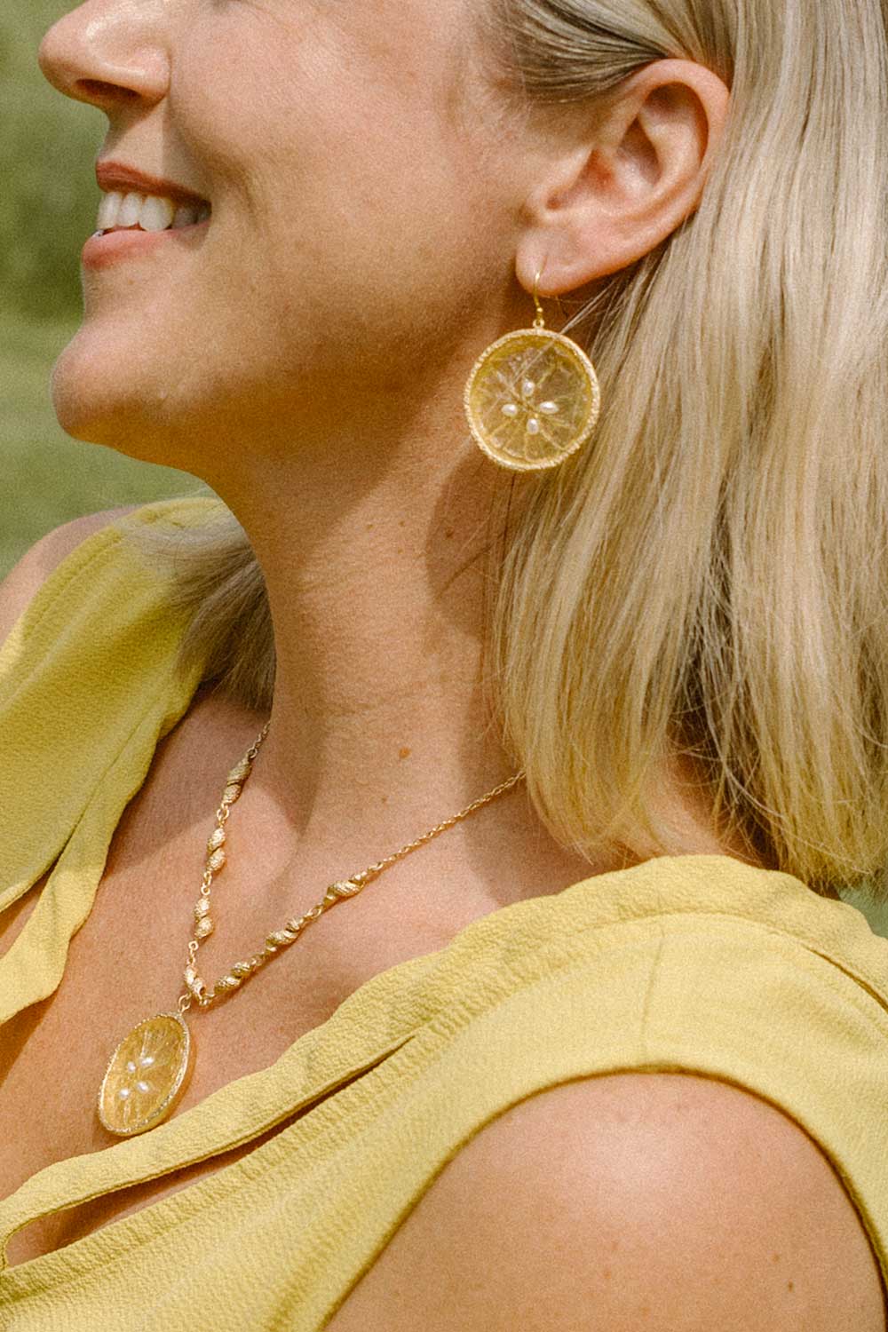 A close-up lifestyle shot of a smiling blonde woman wearing gold-toned circular drop earrings and a matching gold pendant necklace, paired with a yellow top.