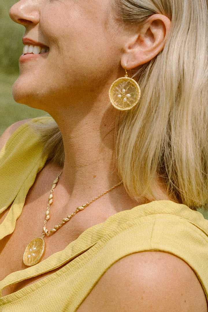 A close-up lifestyle shot of a smiling blonde woman wearing gold-toned circular drop earrings and a matching gold pendant necklace, paired with a yellow top.