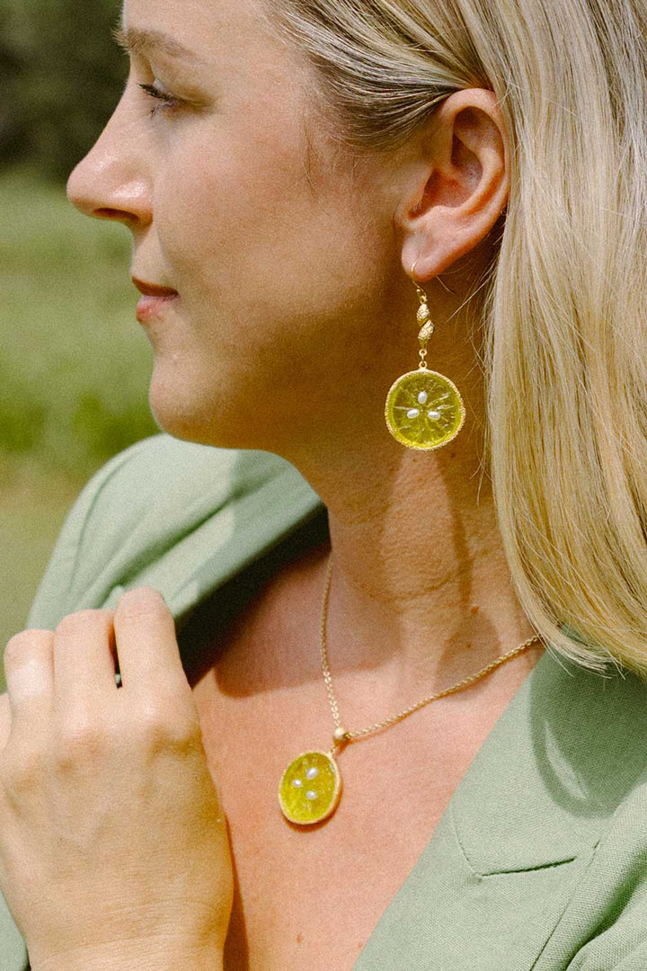 A woman in profile wearing matching gold-toned jewelry, including a necklace and dangling earrings with round yellow-green translucent stone-like pendants, against a softly blurred outdoor background.