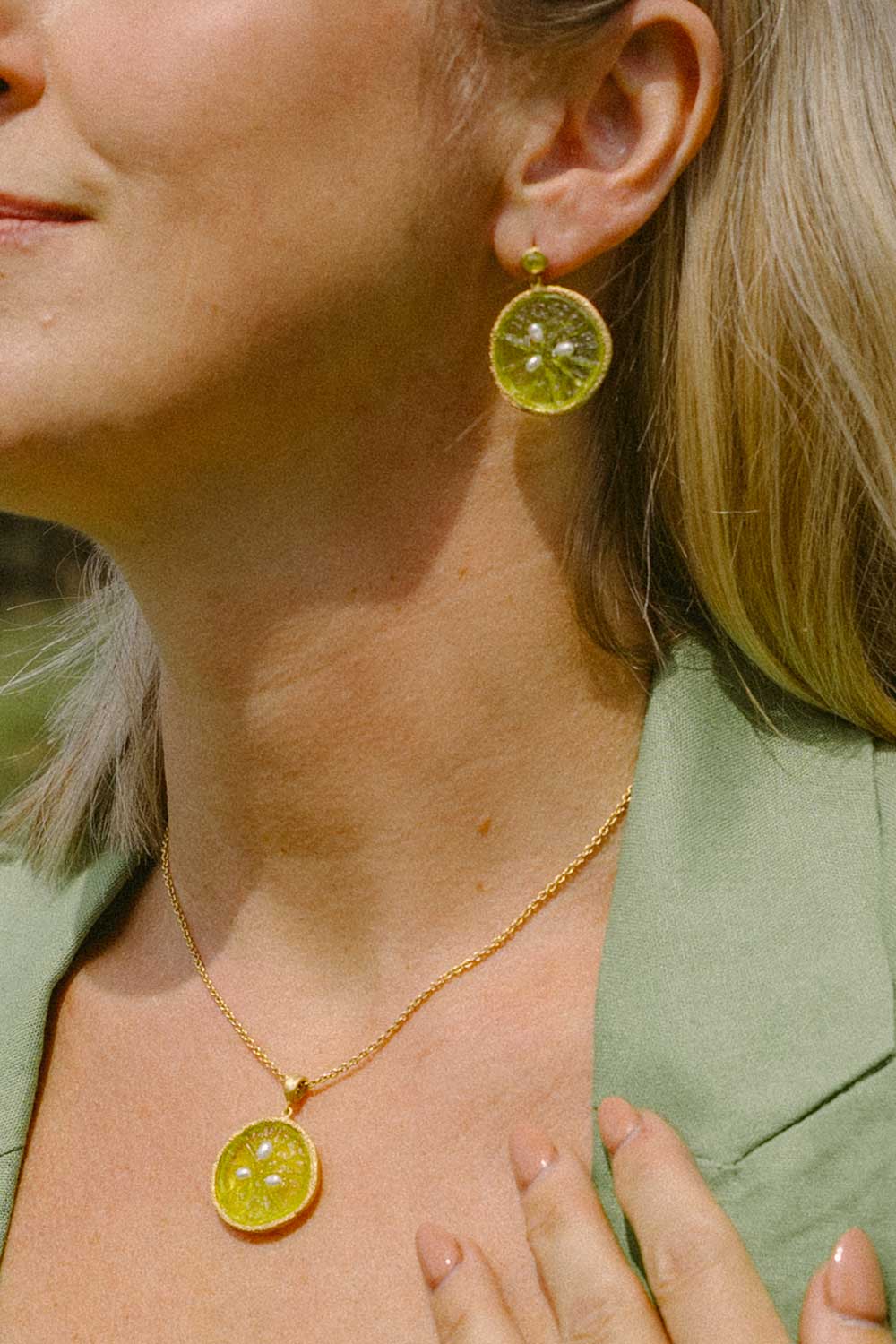 A close-up lifestyle shot of a woman wearing a gold necklace and matching drop earrings, both featuring round yellow-green citrus slice pendants with visible seeds.