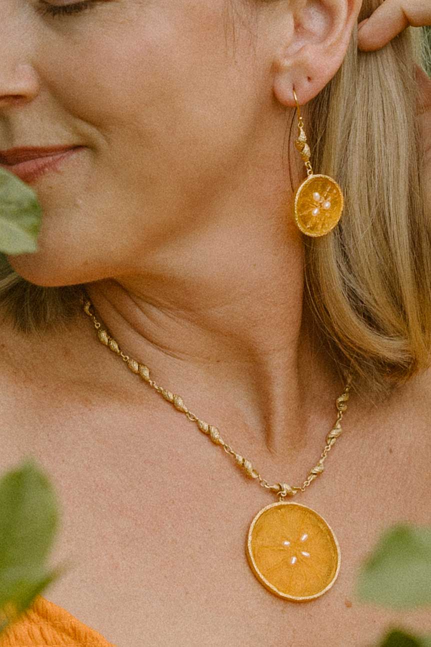 A close-up lifestyle shot of a woman wearing a gold necklace and matching drop earring, both featuring round dried orange slice pendants, with green leaves framing the shot.