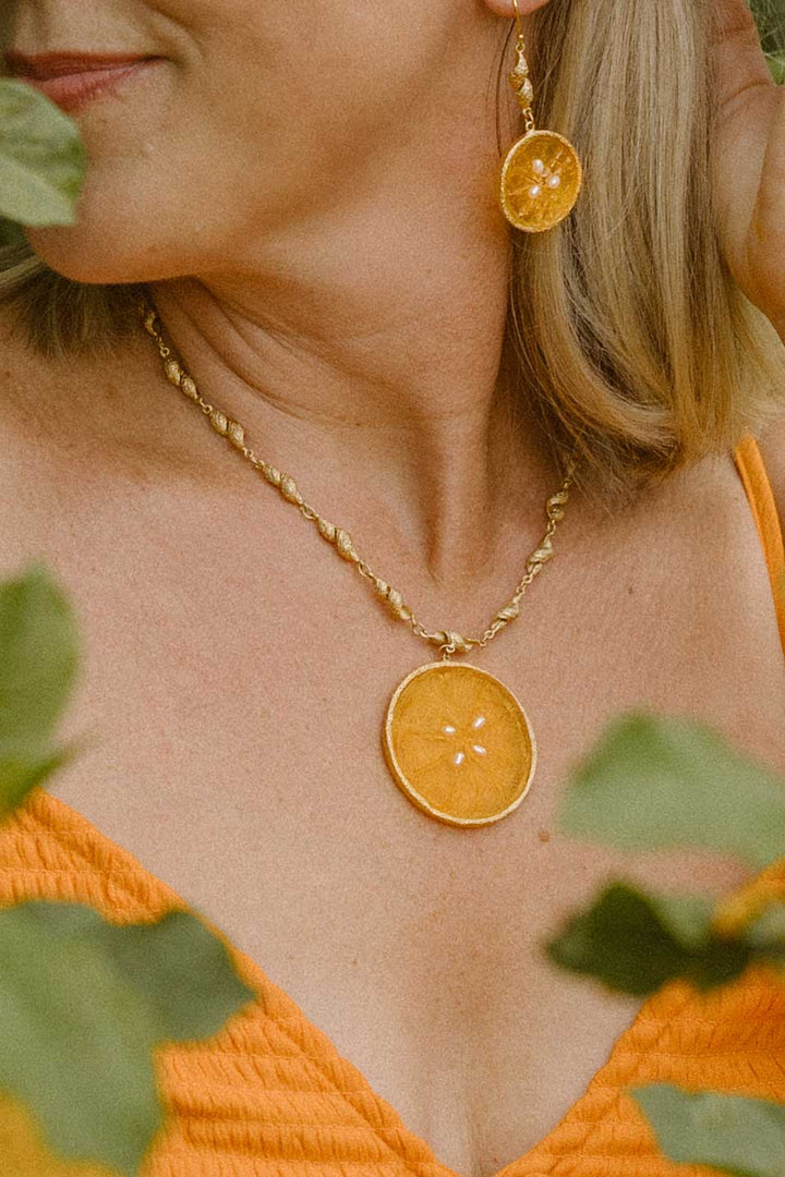 A close-up lifestyle shot of a woman wearing a gold necklace and matching drop earrings, both featuring a circular dried orange slice pendant. She is wearing an orange textured top, with soft green leaves framing the shot.