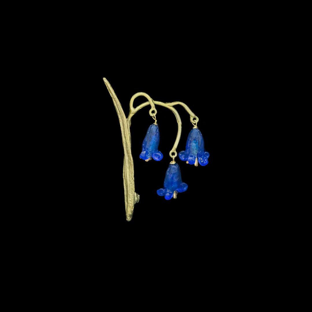 A vintage-style green-toned brooch featuring three dangling blue bell-shaped glass flowers on a curved stem, shown against a solid black background.