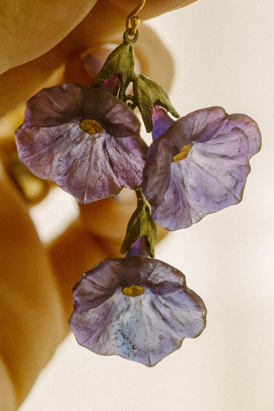 A close-up of a handmade dangle earring featuring cast glass purple and lavender flowers with green sepals and gold-tone hooks, held by a hand against a warm beige background.