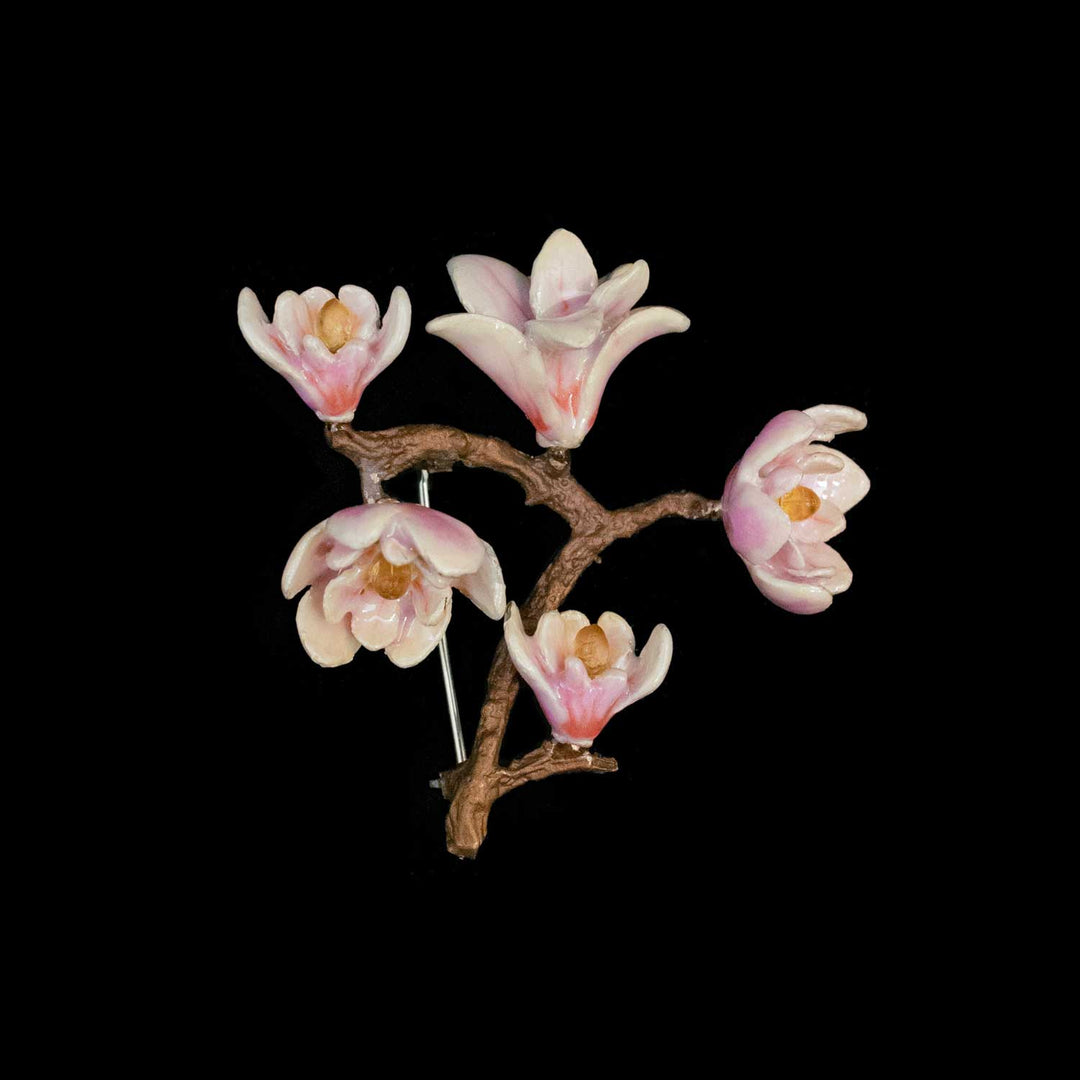 A decorative branch-shaped floral brooch featuring five pale pink and white blossoms with yellow centers on a brown branch, photographed against a solid black background.