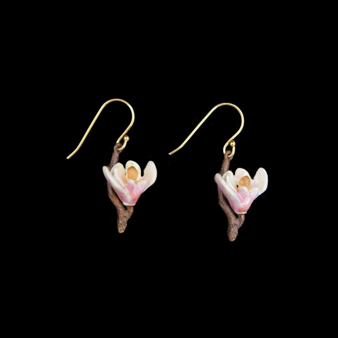 A pair of dangling earrings featuring small white and pink magnolia flowers attached to brown twig-shaped stems, suspended from gold fishhook wires, shown against a solid black background.