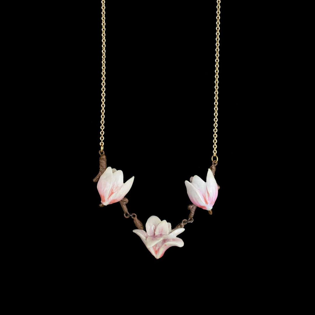 A delicate necklace featuring three sculpted white magnolia blossoms with soft pink edges, attached to a fine gold-toned chain against a solid black background.