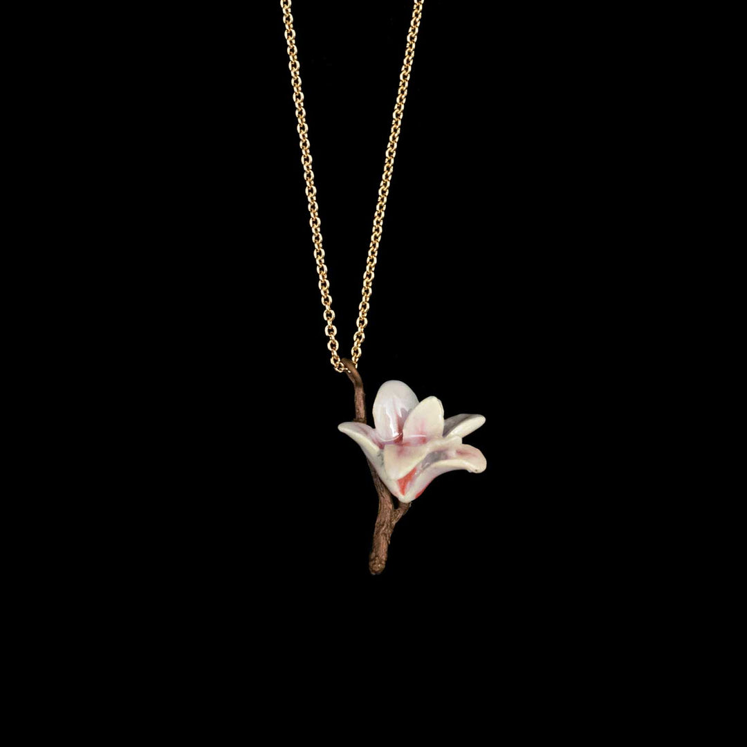 A delicate gold chain necklace featuring a pendant of one white magnolia blossoms with soft pink centers and a brown branch, set against a solid black background.