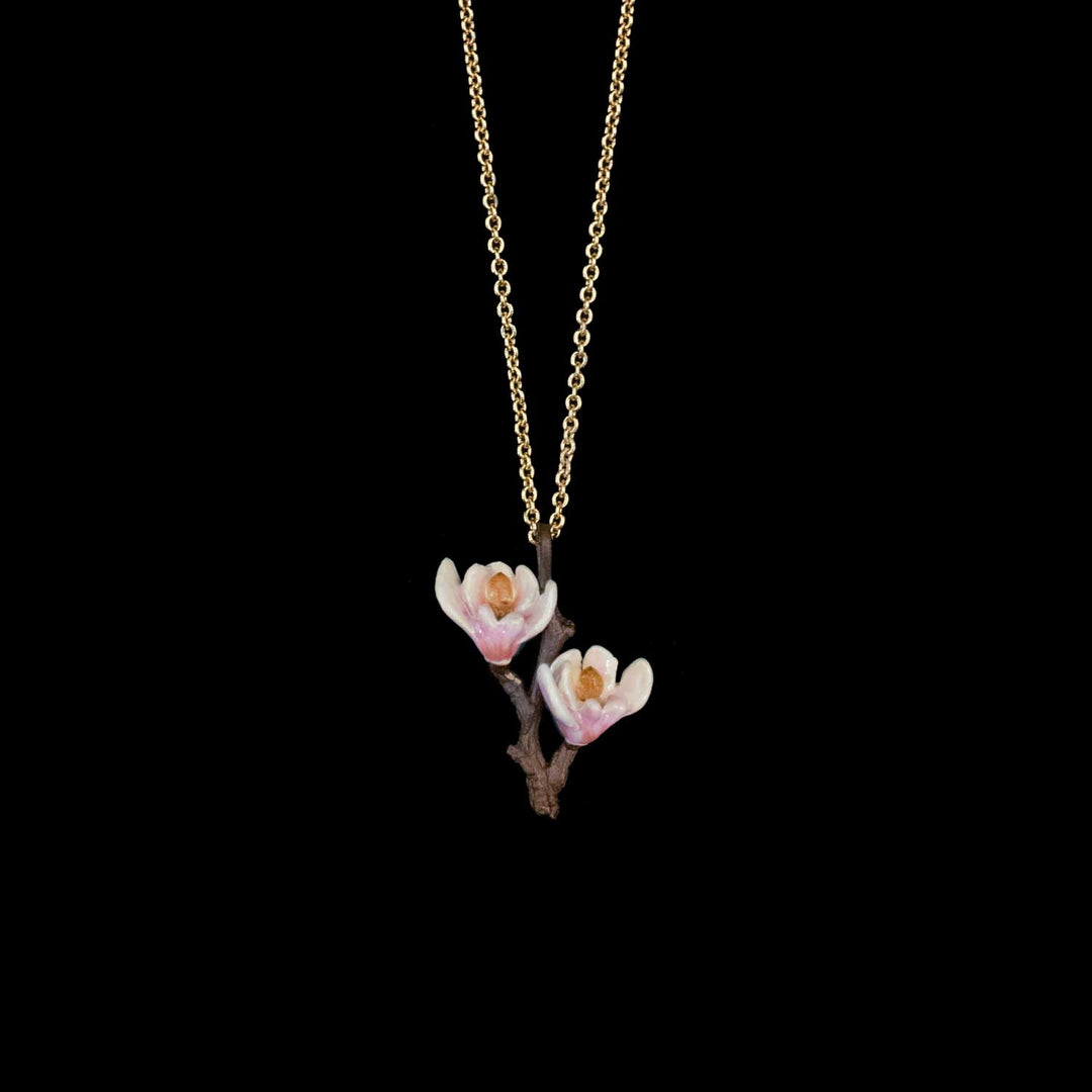 A delicate gold chain necklace featuring a pendant of two white magnolia blossoms with soft pink centers and a brown branch, set against a solid black background.