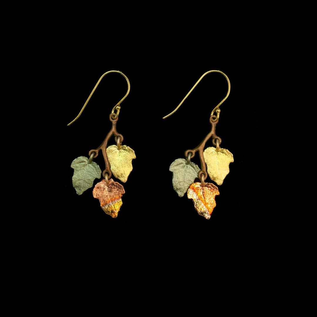 A pair of dangling earrings with gold-tone fishhook wires and small leaf charms in muted green, pale yellow, and copper-orange tones, shown on a solid black background.