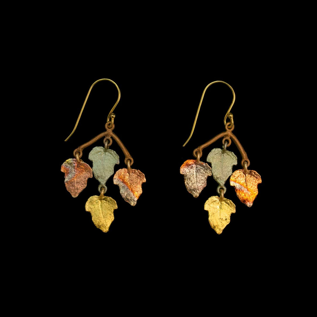 A pair of dangling earrings with gold-tone fishhook wires and branch-shaped tops, each holding three small leaf charms in different metallic colors including gold, silver, bronze, and copper, shown against a solid black background.