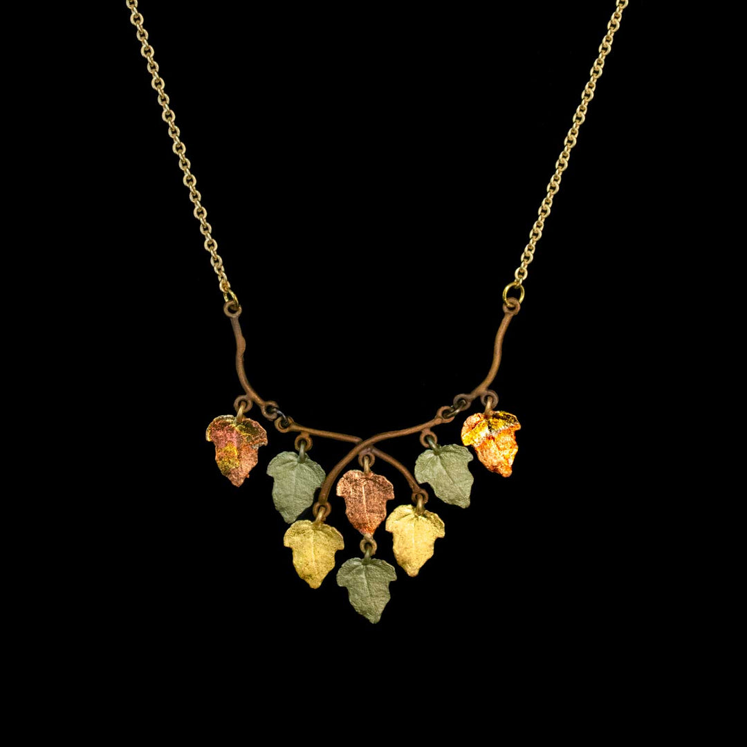 A gold chain necklace featuring a branch-shaped pendant with seven dangling leaves in various colors including green, yellow, orange, and red, set against a solid black background.
