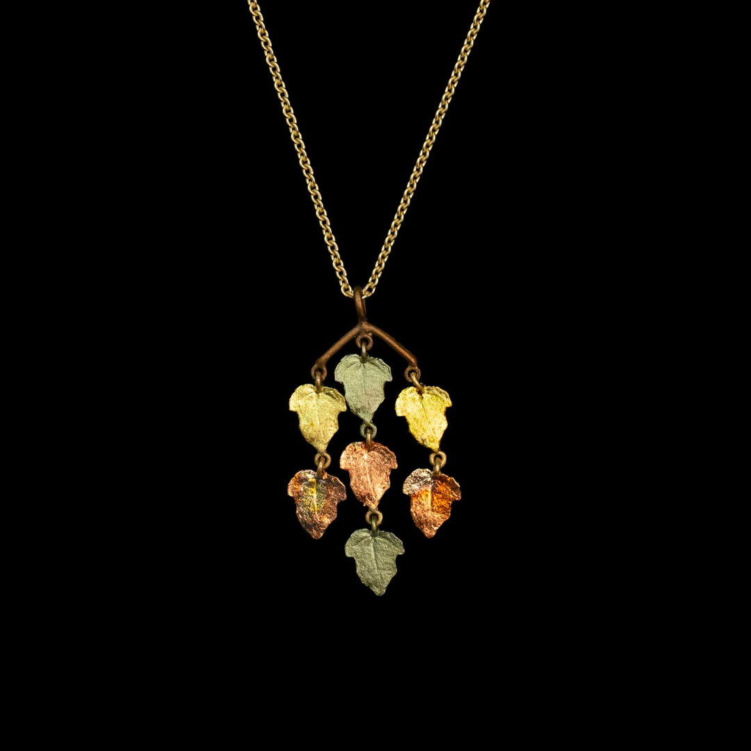 A gold chain necklace with a branch-shaped pendant holding five dangling leaf charms in assorted warm and cool colors, shown against a solid black background.
