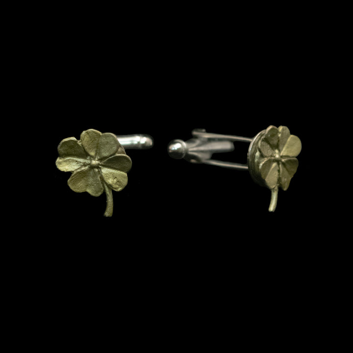A pair of green-toned cufflinks shaped like four-leaf clovers, one shown from the front and the other from the side/back, set against a solid black background.
